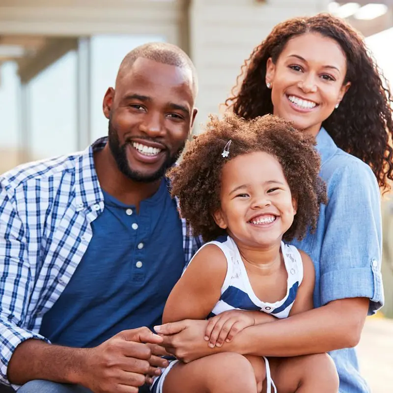 A man, woman, and young child are sitting outside and smiling at the camera. The child is sitting on the woman's lap.