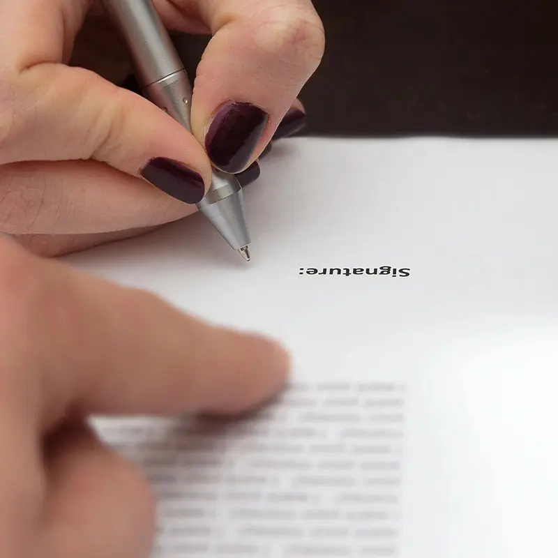 A hand holding a pen is poised to sign a document, while another hand points at the signature line.