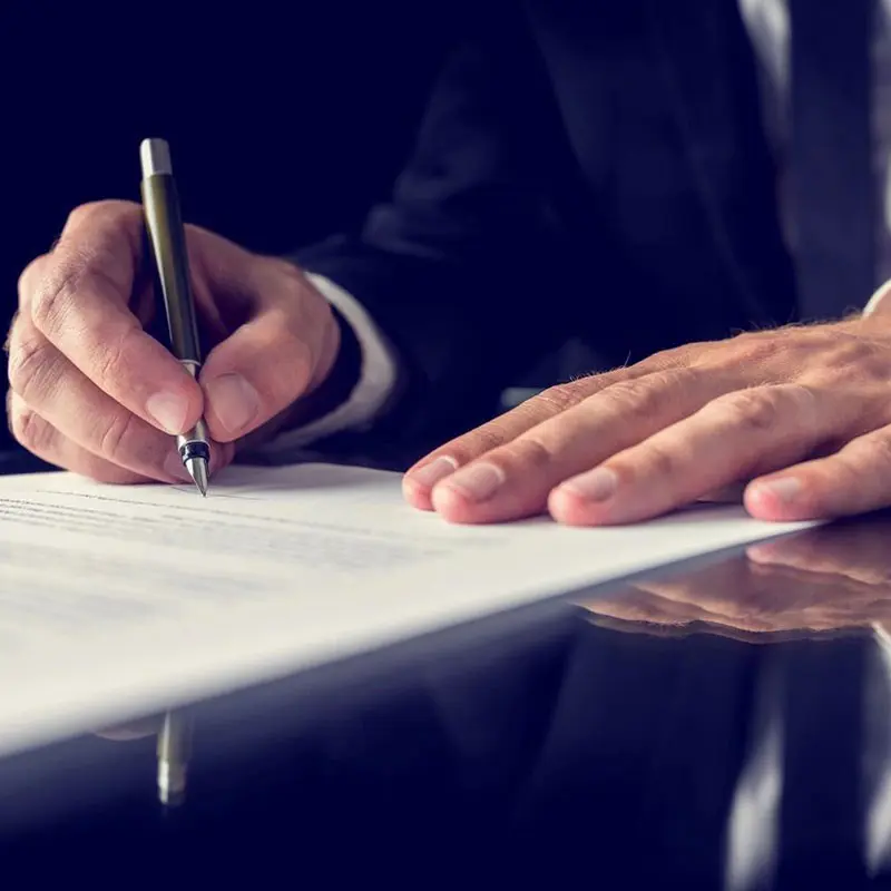 A person in a suit signs a document with a pen on a reflective black surface.